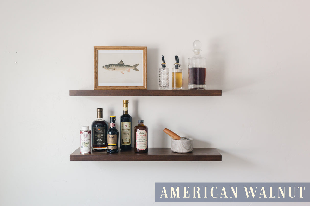 A basswood floating shelf with a American Walnut finish applied decorated with a photo, several spices, and drinks, set against a white wall