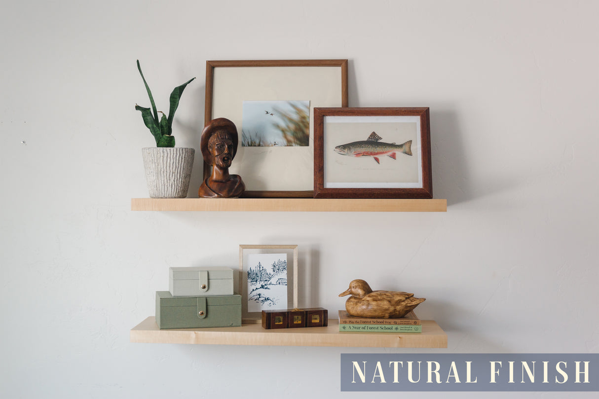 A basswood floating shelf with a natural finish applied decorated with books, little storage containers, wooden mallard, picture frames, and a plant arranged on it, set against a white wall