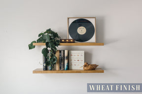 A basswood floating shelf with a wheat finish applied decorated with books, a record, and a plant arranged on it, set against a white wall